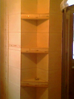 Wnęka narożna z półkami wyłożona jasnymi, kwadratowymi płytkami ceramicznymi w łazience. Widoczne trzy półki z dekoracyjnym obramowaniem. Niska jakość zdjęcia.